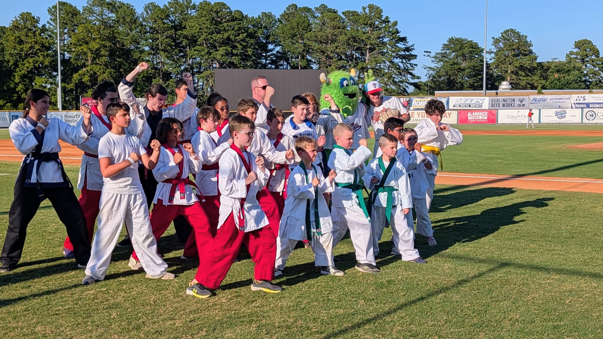 Karate USA students in fighting stance on the outfield grass with the Burlington Sock Puppets mascot cheering behind them.