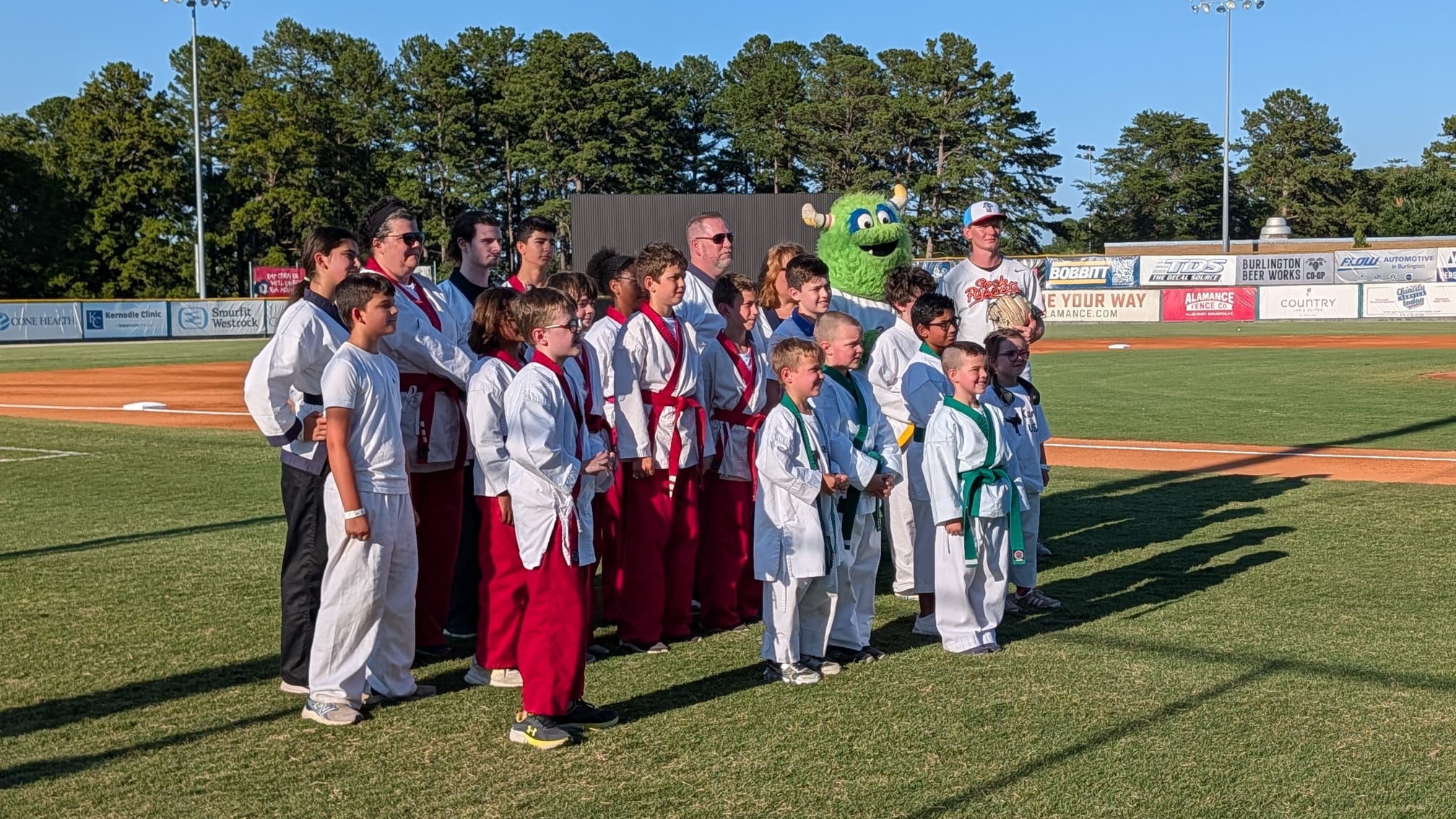 Group photo of the Karate USA demo team with the Sock Puppets mascot and a player at Burlington Athletic Stadium.